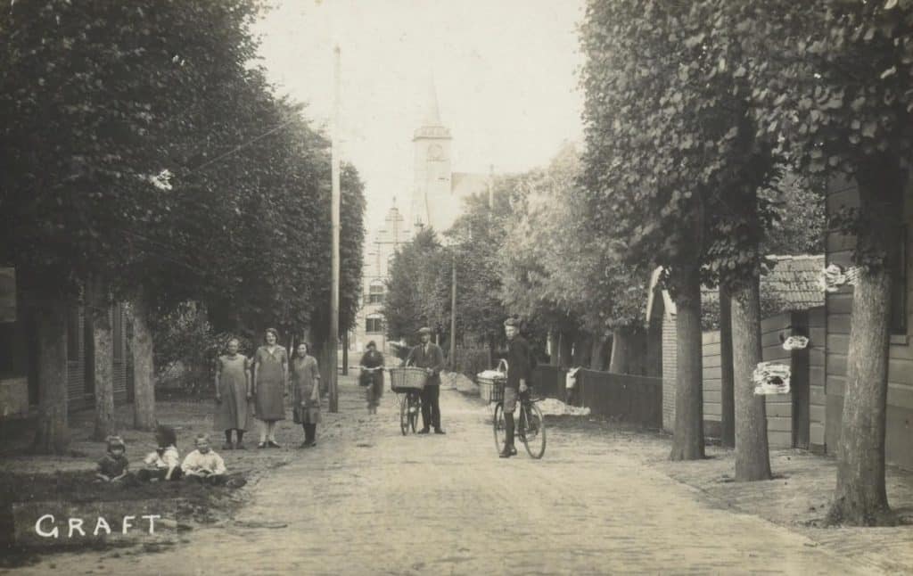 Prentbriefkaart van Raadhuis Graft. Zicht tussen bomen door op het Raadhuis. De fotograaf stond op de hoek van Graft en keek richting raadhuis. Fiets en telefoon hebben hun intrede gedaan, hoedjes en klompen zijn grotendeels verdwenen. Maar de geschoren bomen bepalen nog steeds het straatbeeld. Circa 1927 Collectie Kaptein.